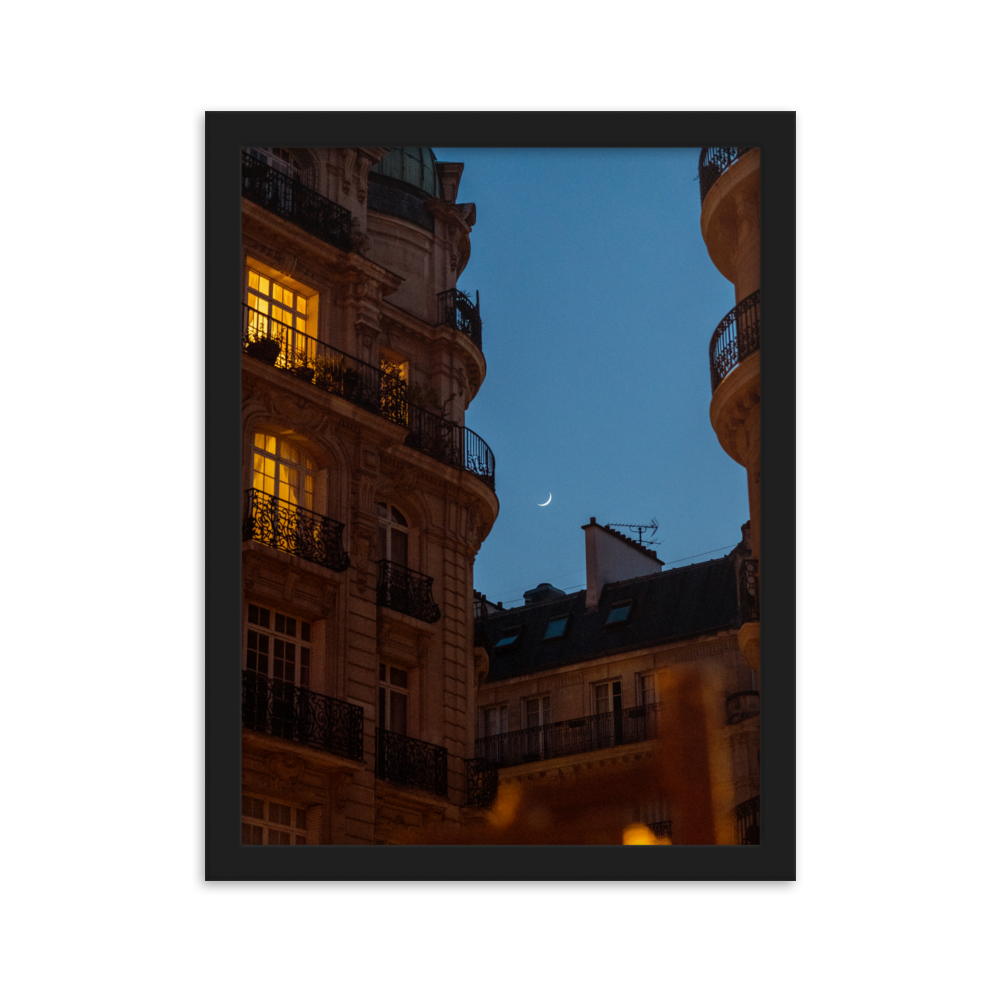 Croissant de lune entre des immeubles haussmanniens éclairés la nuit.