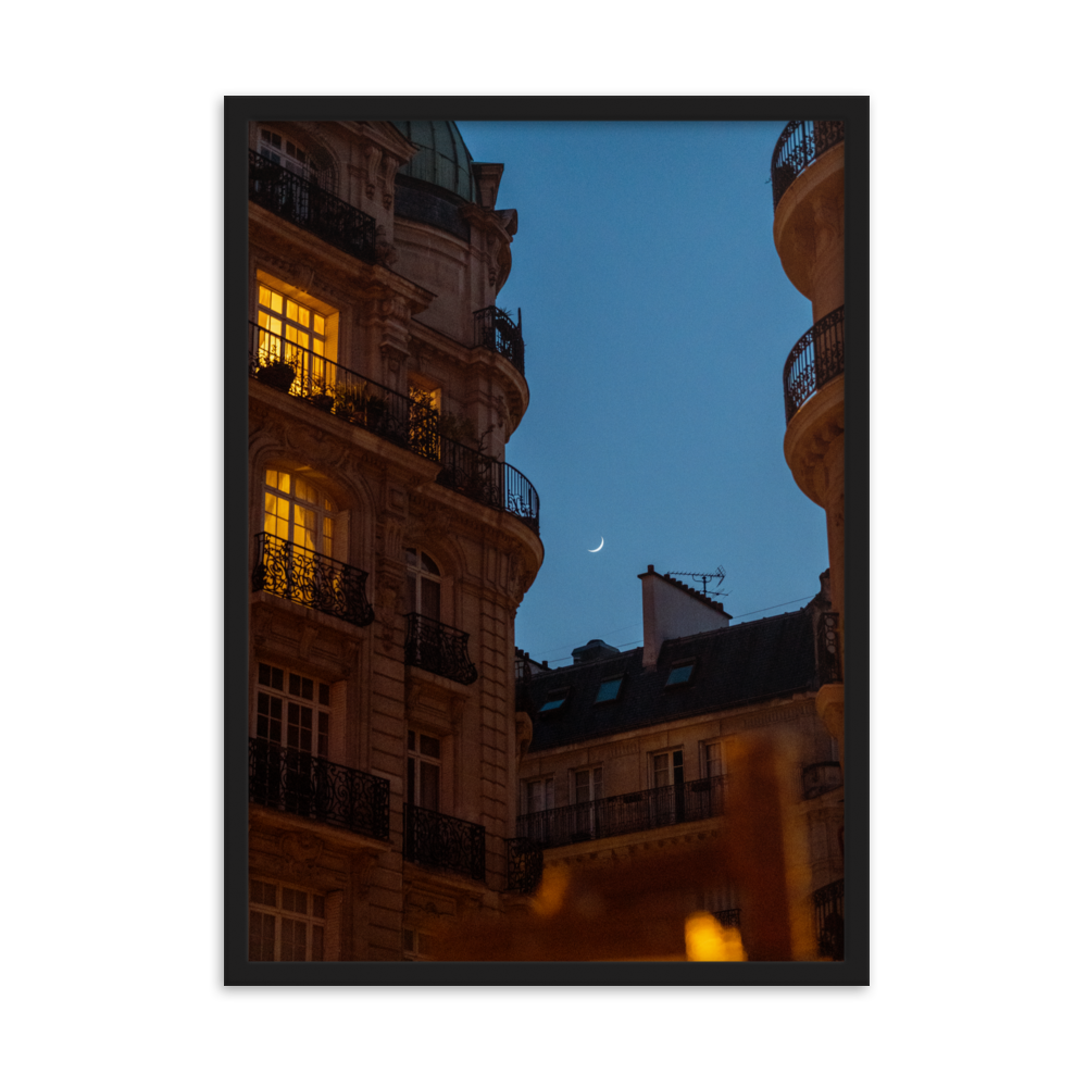 Croissant de lune entre des immeubles haussmanniens éclairés la nuit.