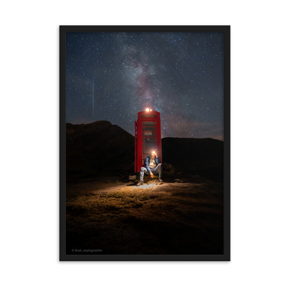 Cabine téléphonique rouge illuminée au milieu de montagnes sombres sous un ciel étoilé par la Voie Lactée, un homme contemple la scène – œuvre signée Brandon Valette.