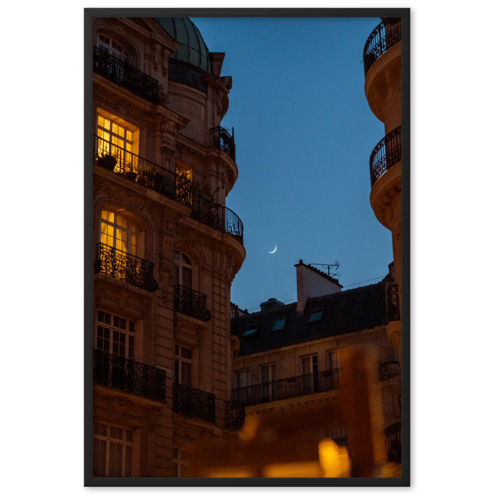 Croissant de lune entre des immeubles haussmanniens éclairés la nuit.