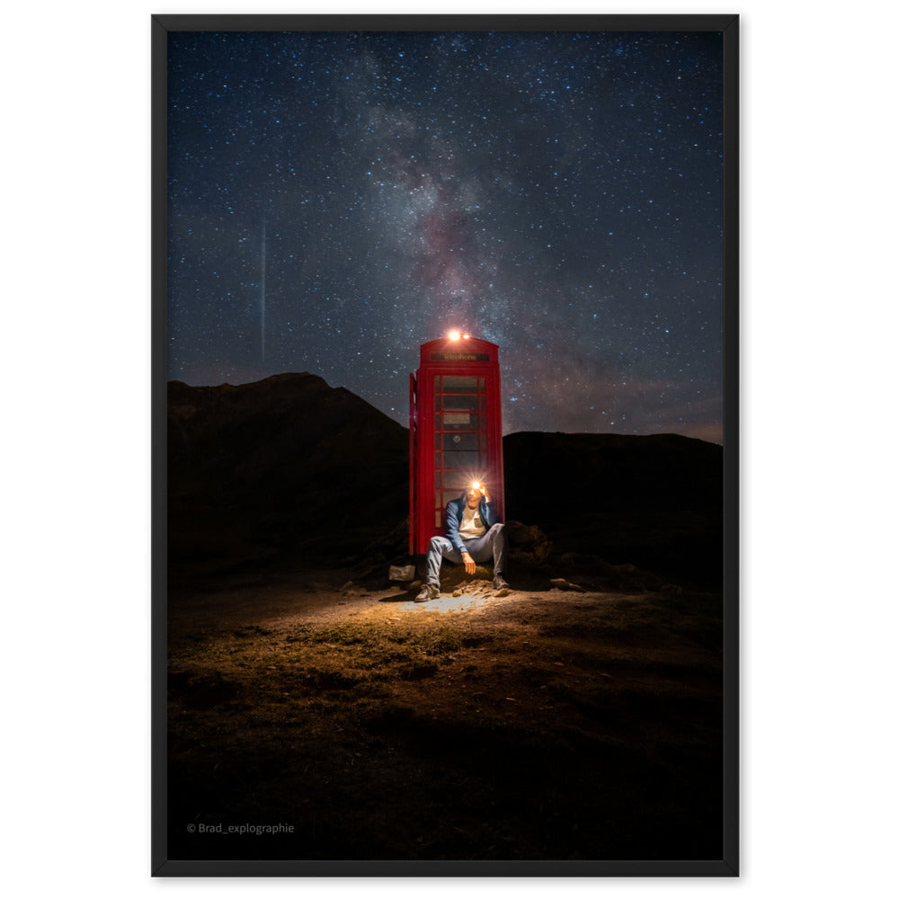 Cabine téléphonique rouge illuminée au milieu de montagnes sombres sous un ciel étoilé par la Voie Lactée, un homme contemple la scène – œuvre signée Brandon Valette.