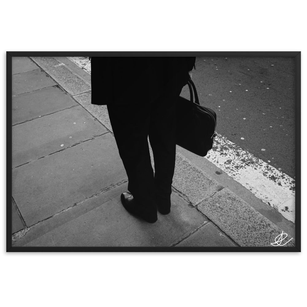 Photographie noir et blanc 'L'Homme d'Affaires' par Ilan Shoham, capturant une scène urbaine quotidienne avec un focus sur une paire de jambes, un sac à documents et un costume noir contre le décor brut du trottoir, encadrée élégamment pour un impact visuel puissant.