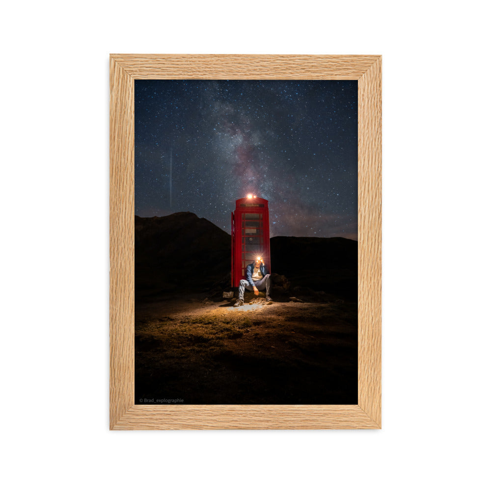 Cabine téléphonique rouge illuminée au milieu de montagnes sombres sous un ciel étoilé par la Voie Lactée, un homme contemple la scène – œuvre signée Brandon Valette.