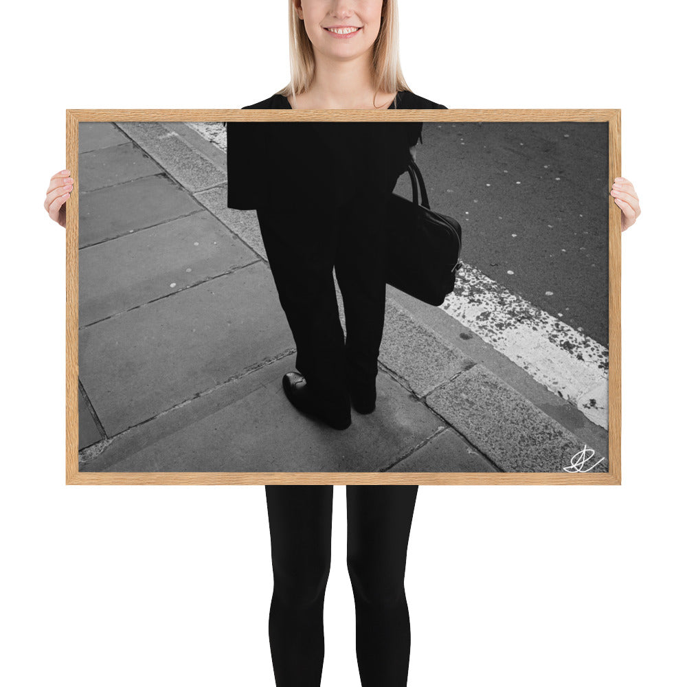 Photographie noir et blanc 'L'Homme d'Affaires' par Ilan Shoham, capturant une scène urbaine quotidienne avec un focus sur une paire de jambes, un sac à documents et un costume noir contre le décor brut du trottoir, encadrée élégamment pour un impact visuel puissant.