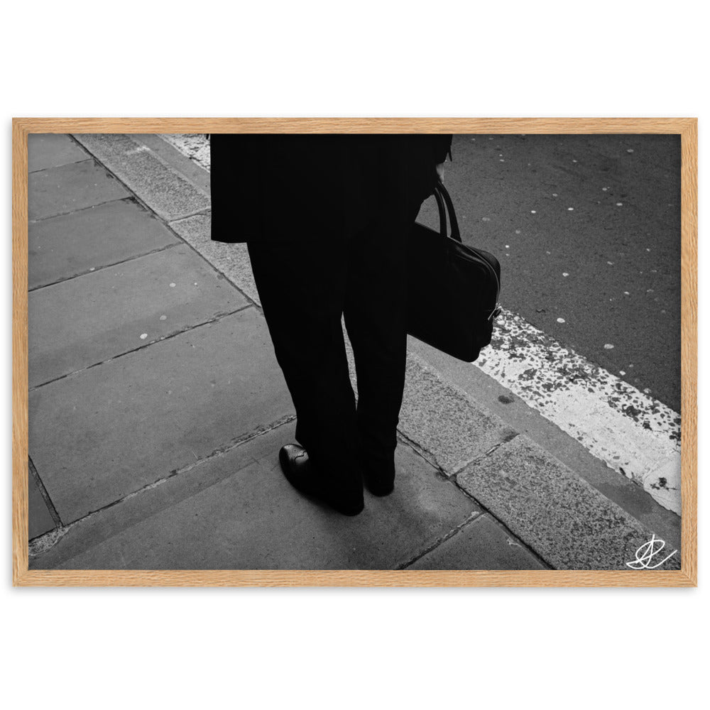 Photographie noir et blanc 'L'Homme d'Affaires' par Ilan Shoham, capturant une scène urbaine quotidienne avec un focus sur une paire de jambes, un sac à documents et un costume noir contre le décor brut du trottoir, encadrée élégamment pour un impact visuel puissant.