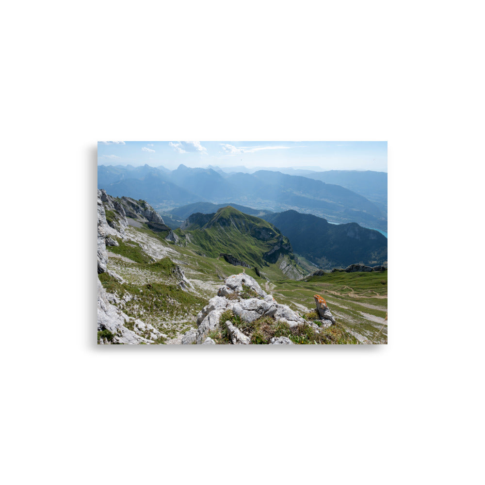 Vue panoramique du lac Annecy depuis les hauteurs, entouré de montagnes majestueuses avec un ciel serein au-dessus. Une représentation fidèle de la beauté naturelle des Alpes.