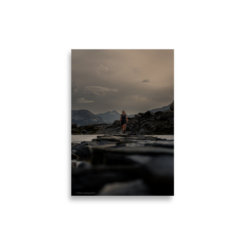 Randonneuse traversant un lac séparé par un chemin de pierres, avec une toile de fond montagneuse, œuvre de Brad_explographie.