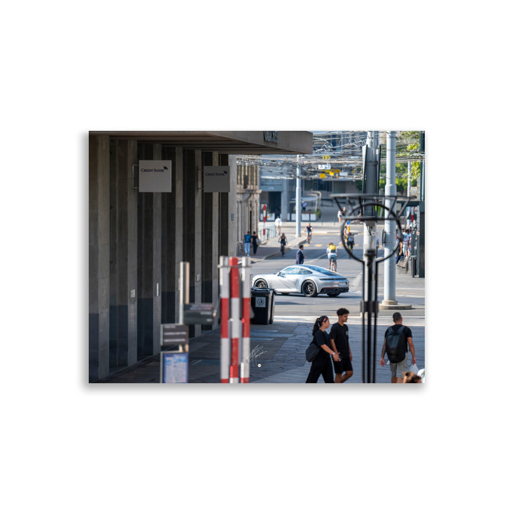 Photographie urbaine montrant la façade de la banque Crédit Suisse à Genève, avec des passants, des cyclistes, et une élégante Porsche 911 grise en mouvement au premier plan.