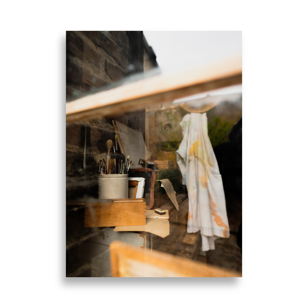 Poster d'un vieil atelier artistique vu à travers une vitre, avec des pinceaux, des boîtes en bois, et une chemise tachée de peinture