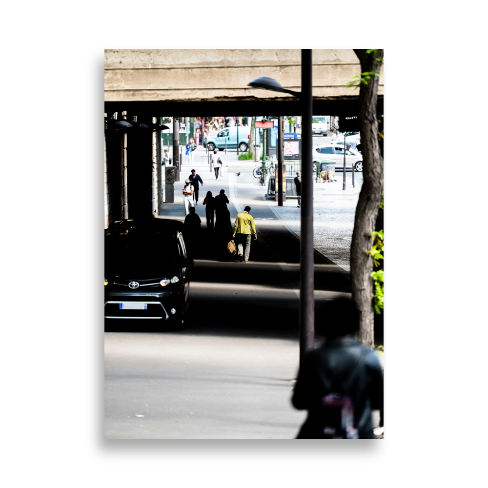 Un homme en veste verte kaki sous un pont ensoleillé.