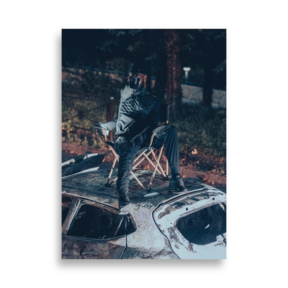 Photographie artistique 'Le Maestro' - Un rappeur captivant brandit une arme factice sur une voiture incendiée, symbolisant la puissance de sa musique.