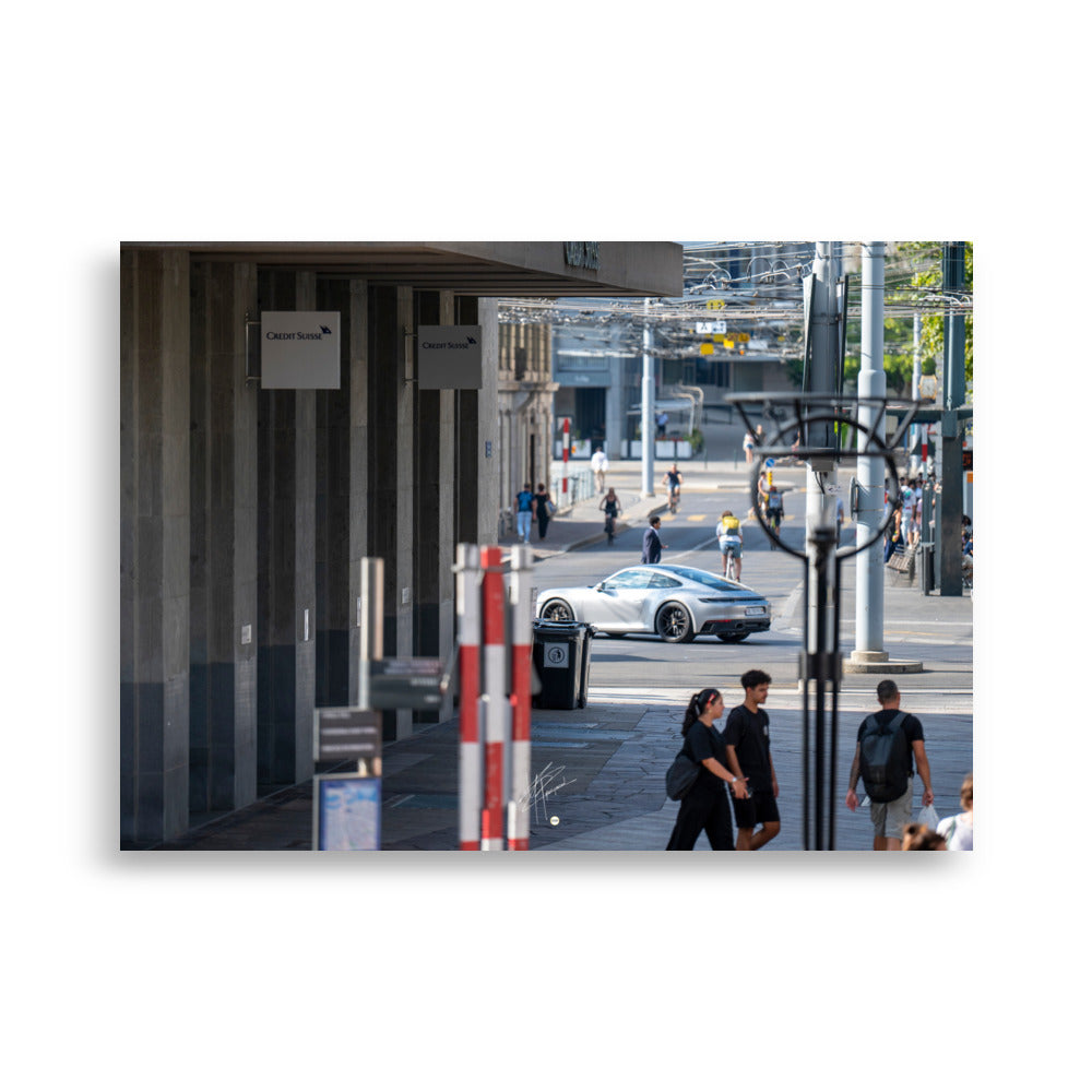 Photographie urbaine montrant la façade de la banque Crédit Suisse à Genève, avec des passants, des cyclistes, et une élégante Porsche 911 grise en mouvement au premier plan.