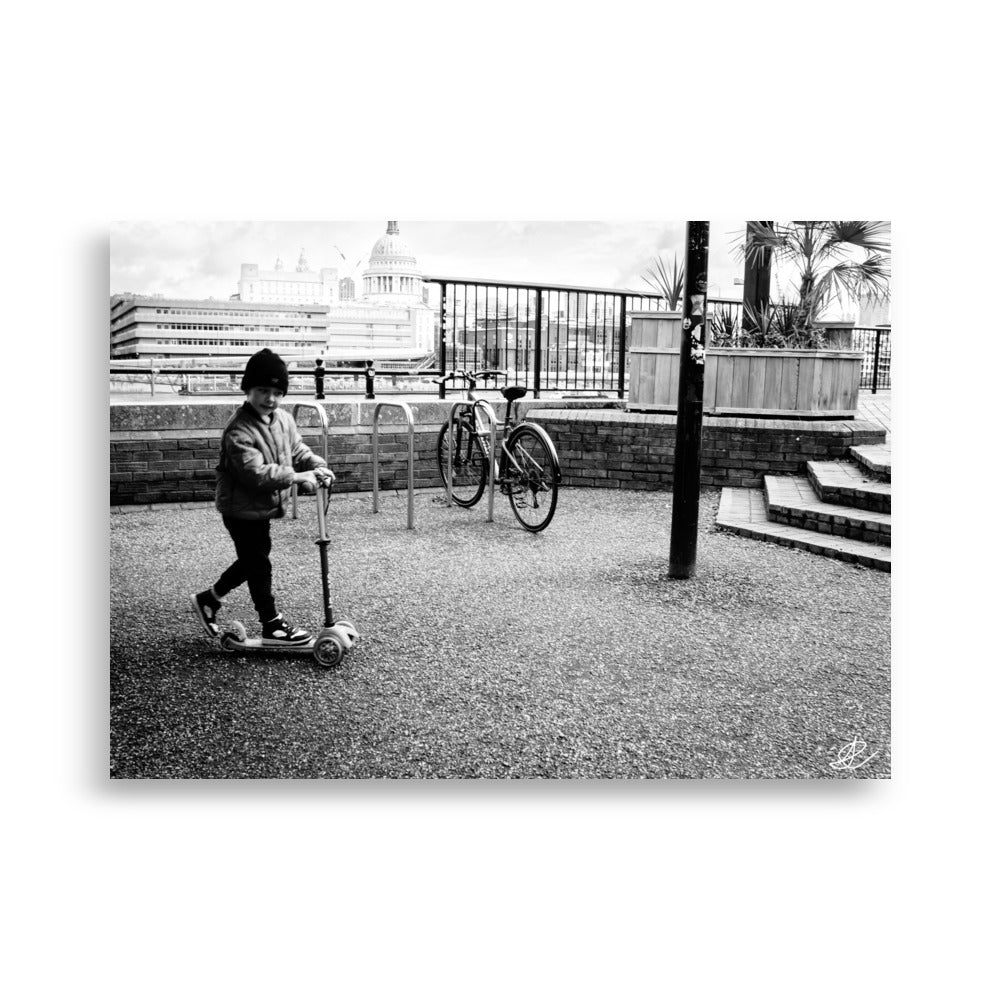 Photographie noir et blanc 'Trottinette' par Ilan Shoham, représentant un enfant joyeux trottinant sur les pavés de Londres, symbolisant l'innocence et la liberté