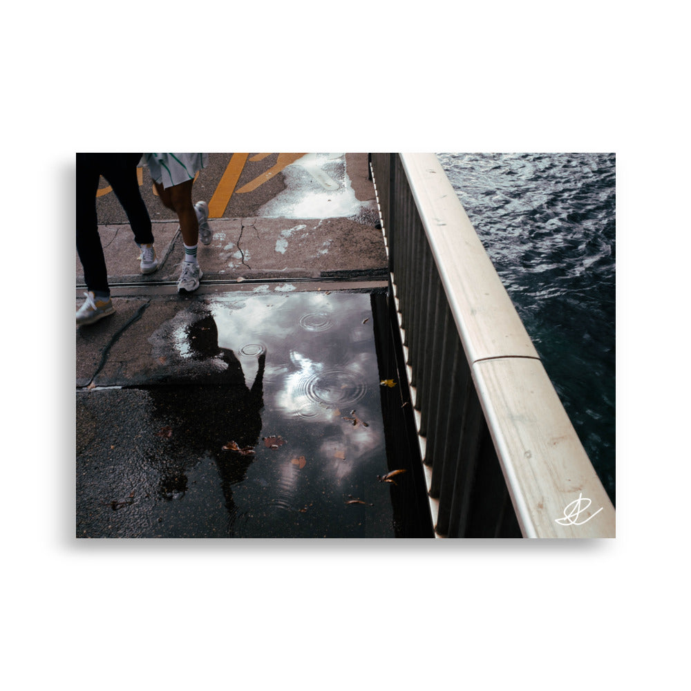 Photographie de rue capturant le reflet de deux passants dans une flaque, par Ilan Shoham, offrant une perspective unique et surréaliste de la vie urbaine quotidienne.