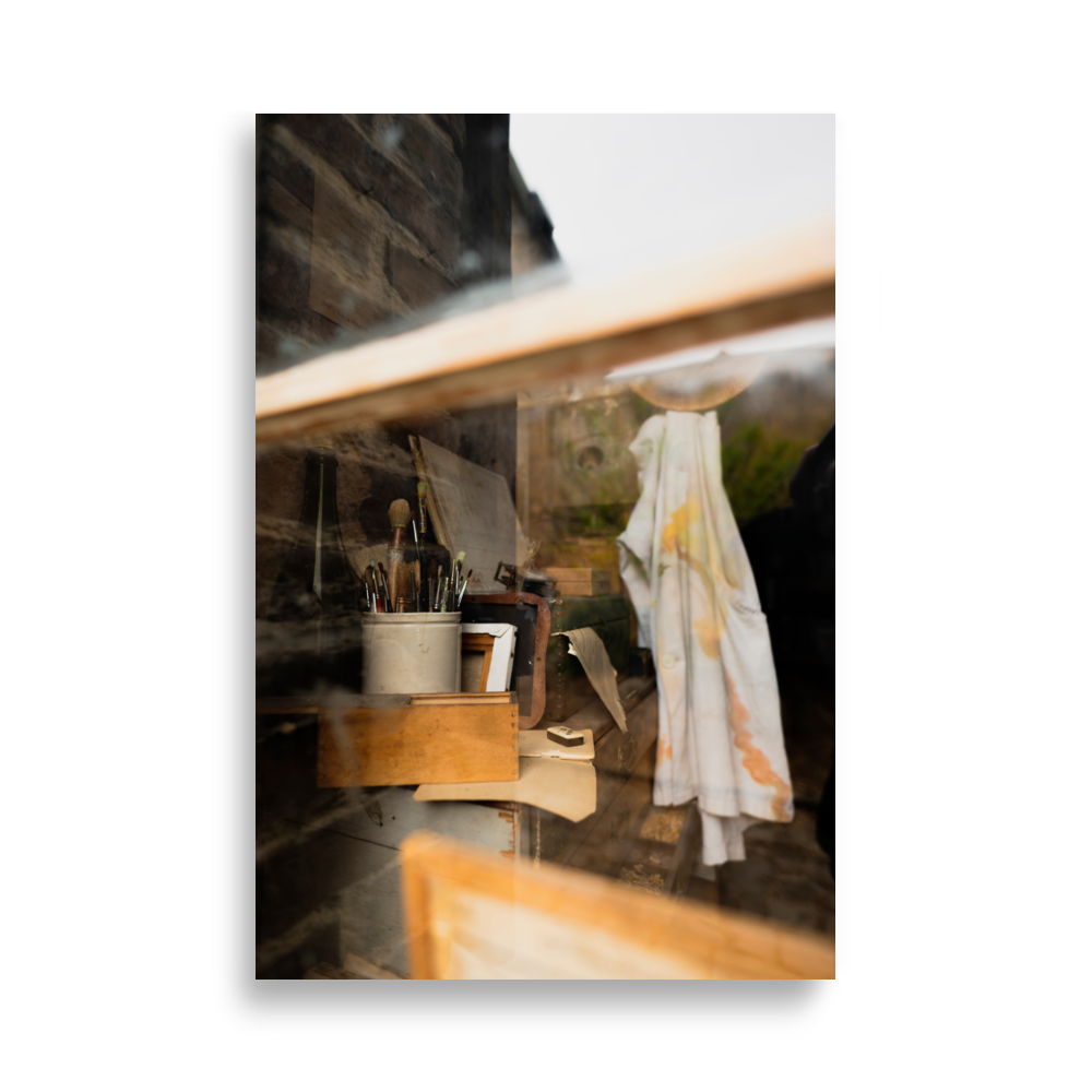 Poster d'un vieil atelier artistique vu à travers une vitre, avec des pinceaux, des boîtes en bois, et une chemise tachée de peinture