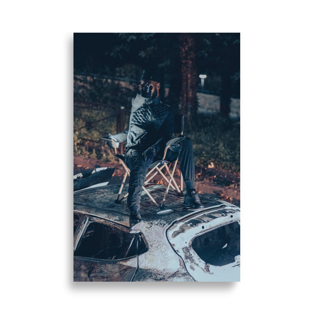 Photographie artistique 'Le Maestro' - Un rappeur captivant brandit une arme factice sur une voiture incendiée, symbolisant la puissance de sa musique.