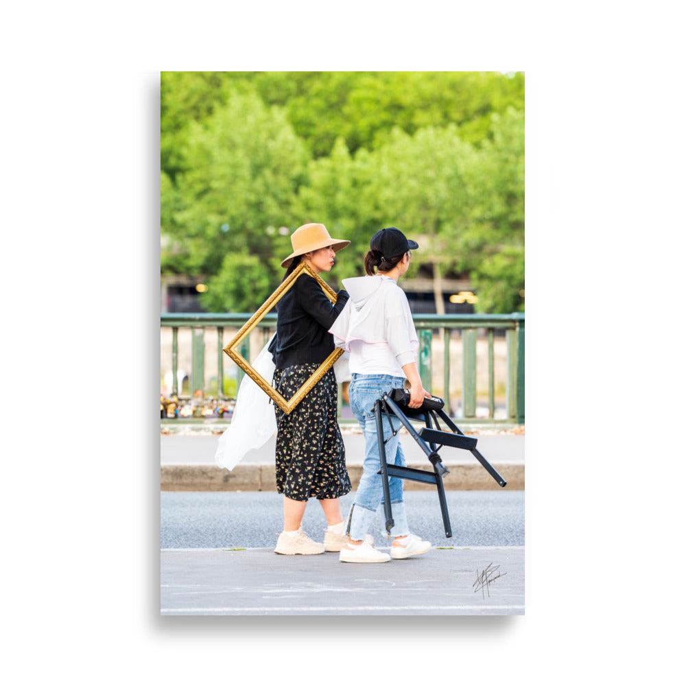 Photographie de deux femmes dans une rue, l'une portant élégamment un cadre en bois, évoquant mystère et art dans le quotidien.