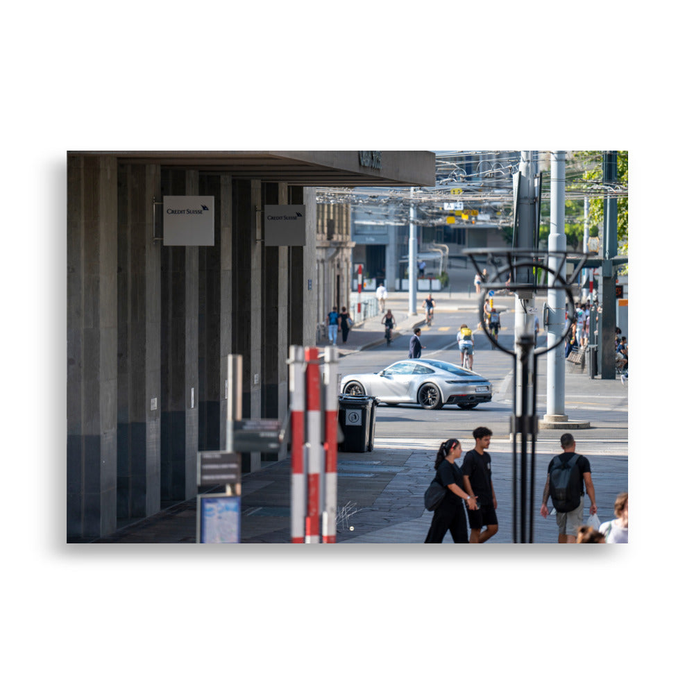 Photographie urbaine montrant la façade de la banque Crédit Suisse à Genève, avec des passants, des cyclistes, et une élégante Porsche 911 grise en mouvement au premier plan.