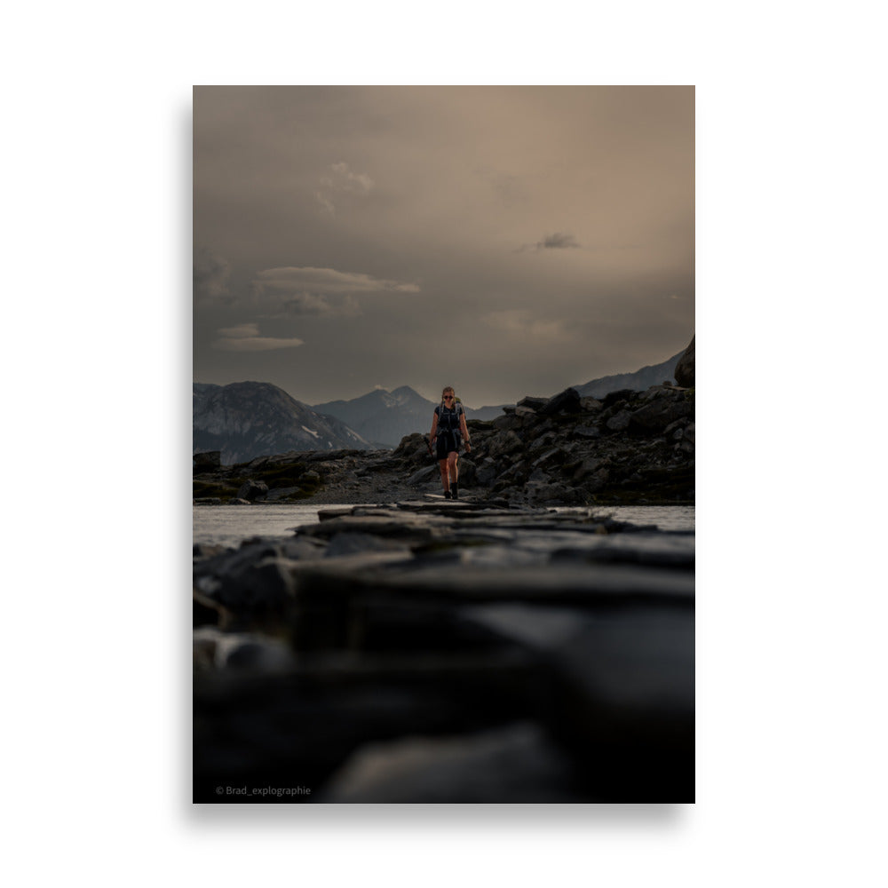 Randonneuse traversant un lac séparé par un chemin de pierres, avec une toile de fond montagneuse, œuvre de Brad_explographie.