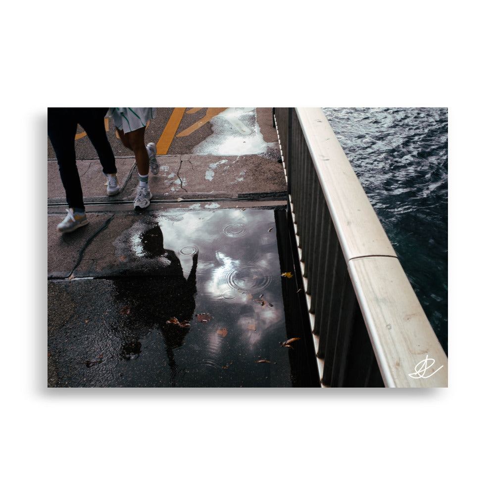 Photographie de rue capturant le reflet de deux passants dans une flaque, par Ilan Shoham, offrant une perspective unique et surréaliste de la vie urbaine quotidienne.