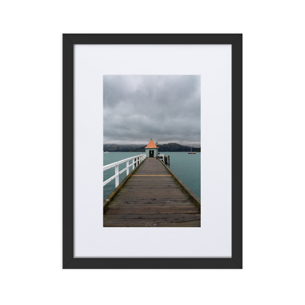 Photographie 'Akaroa NZ' de Charles Coley, offrant une vue sereine sur un lac néo-zélandais avec un petit abri au bout d'un ponton rustique, contrastant délicatement avec un arrière-plan de montagnes majestueuses et un ciel nuageux, ajoutant une dimension de tranquillité et d'évasion à votre espace.
